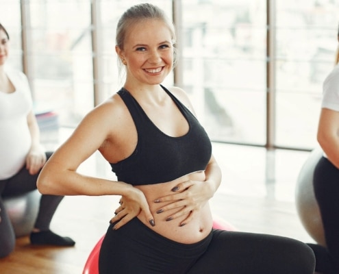 Schwangere Frau sitzt auf Gymnastikball und macht Sport. Sie greift sich dabei an den Babybauch. optimumtraining.at